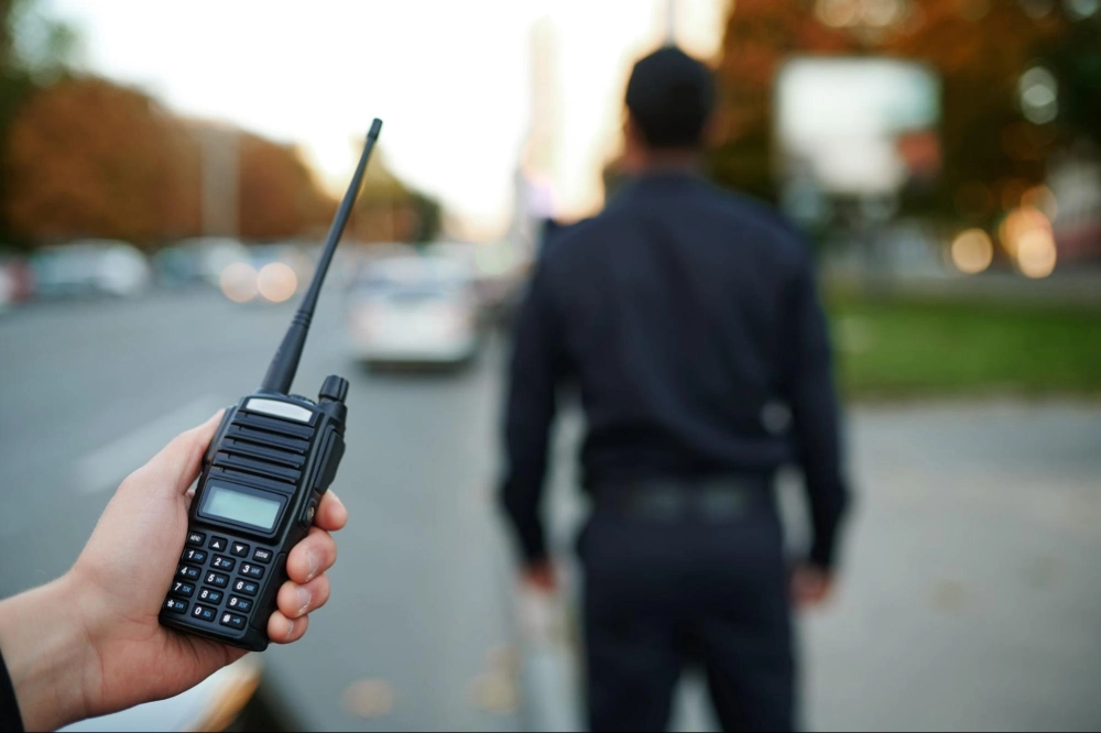 Hand holding a two-way radio with a security guard patrolling in the background, illustrating radio communication training from Guard Training.
