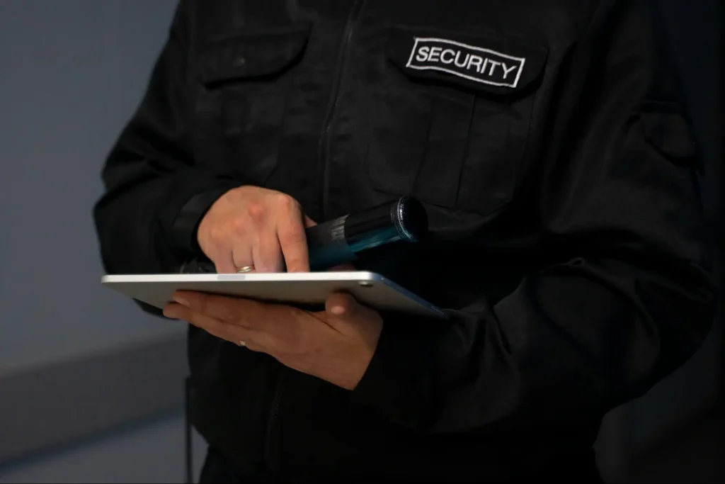 Security guard in black uniform holding a flashlight and using a tablet for digital patrol reporting and security monitoring.