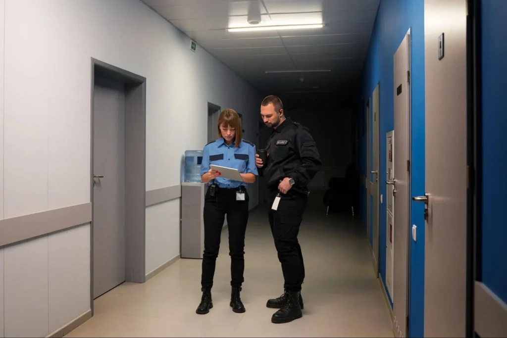 Two security guards in uniform reviewing information on a tablet while coordinating indoor patrol duties in a building hallway.